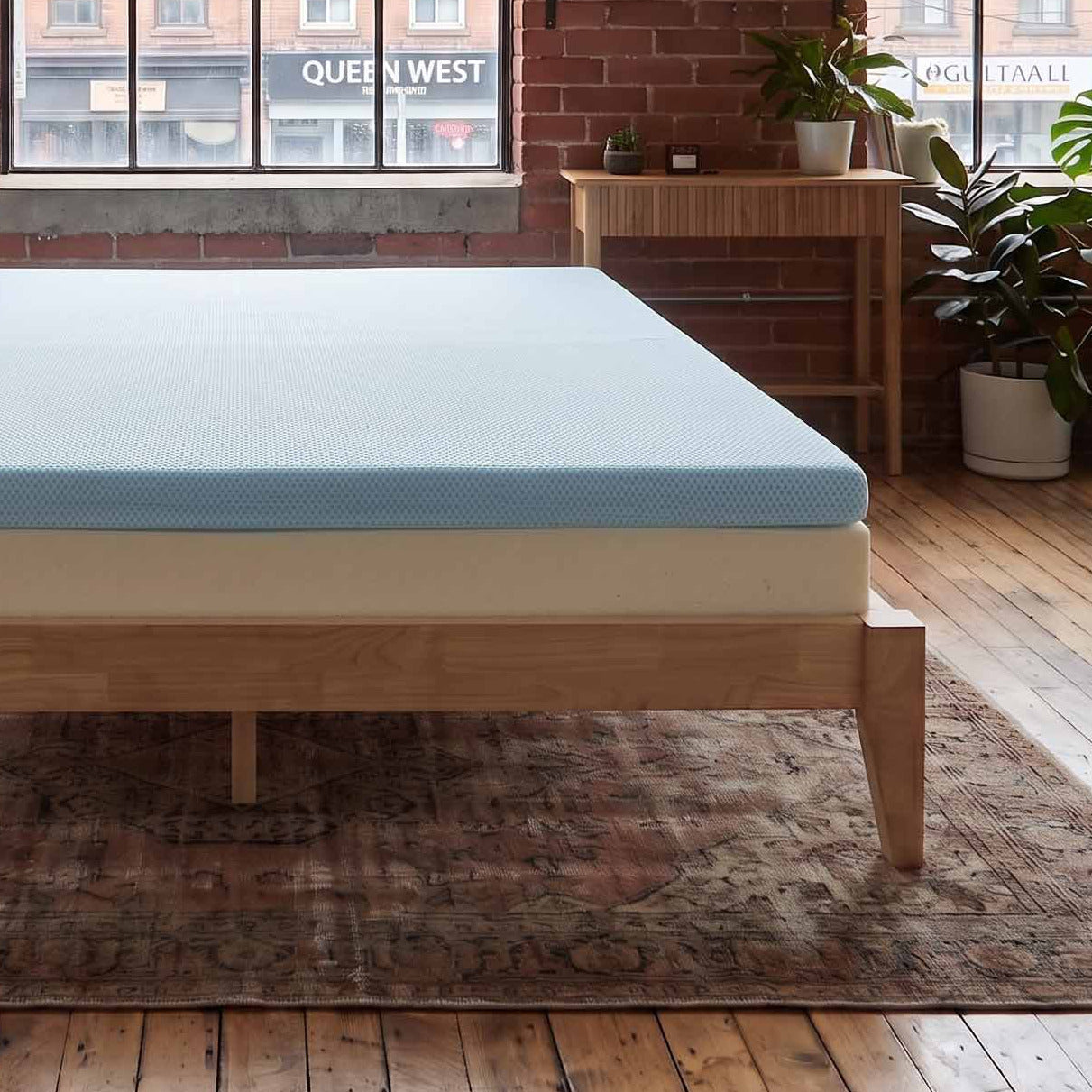 A light blue mattress on a wooden bed frame in a loft-style bedroom with brick walls, large windows, and a patterned rug.