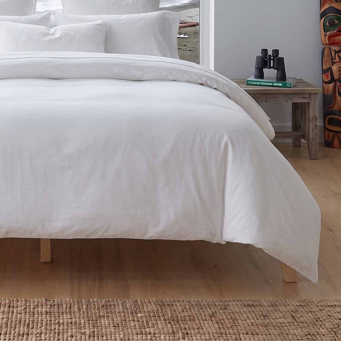 A neatly made bed with white bedding and multiple white pillows sits in a bright bedroom with large windows overlooking an ocean view with rocky islands and mountains. A small wooden side table with books and a plant is visible to the right.