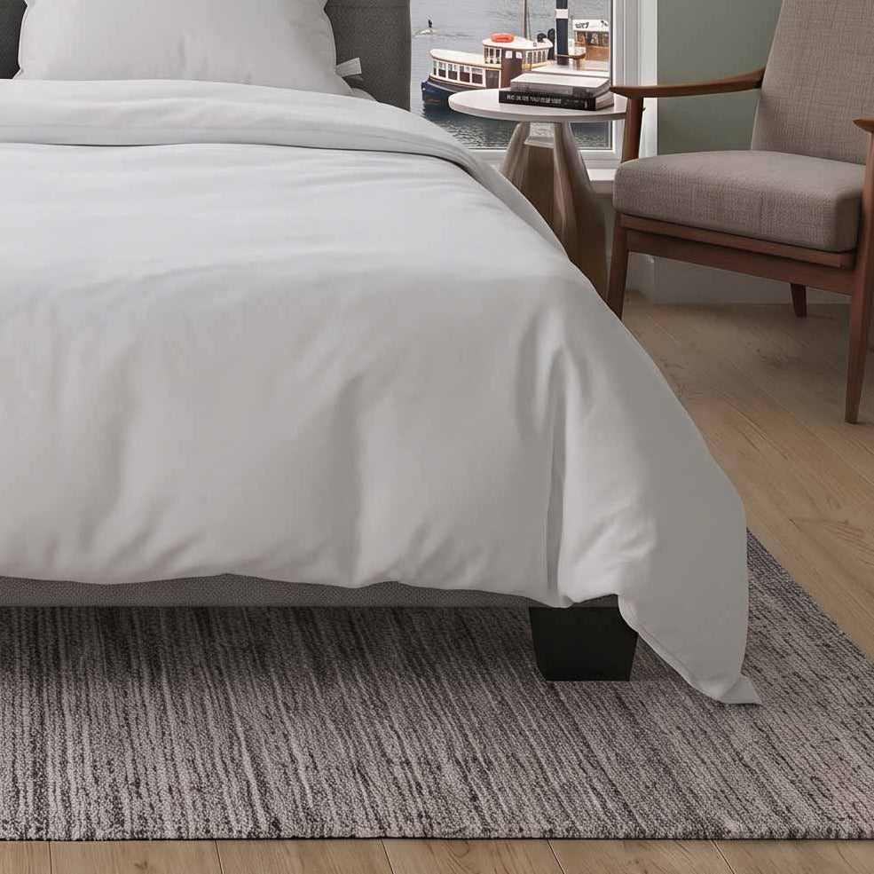 A neatly made bed with white bedding and two white pillows sits in a modern bedroom with a large window overlooking a city and water view. The room includes a gray upholstered headboard, gray rug, side table, chair, and potted plant.