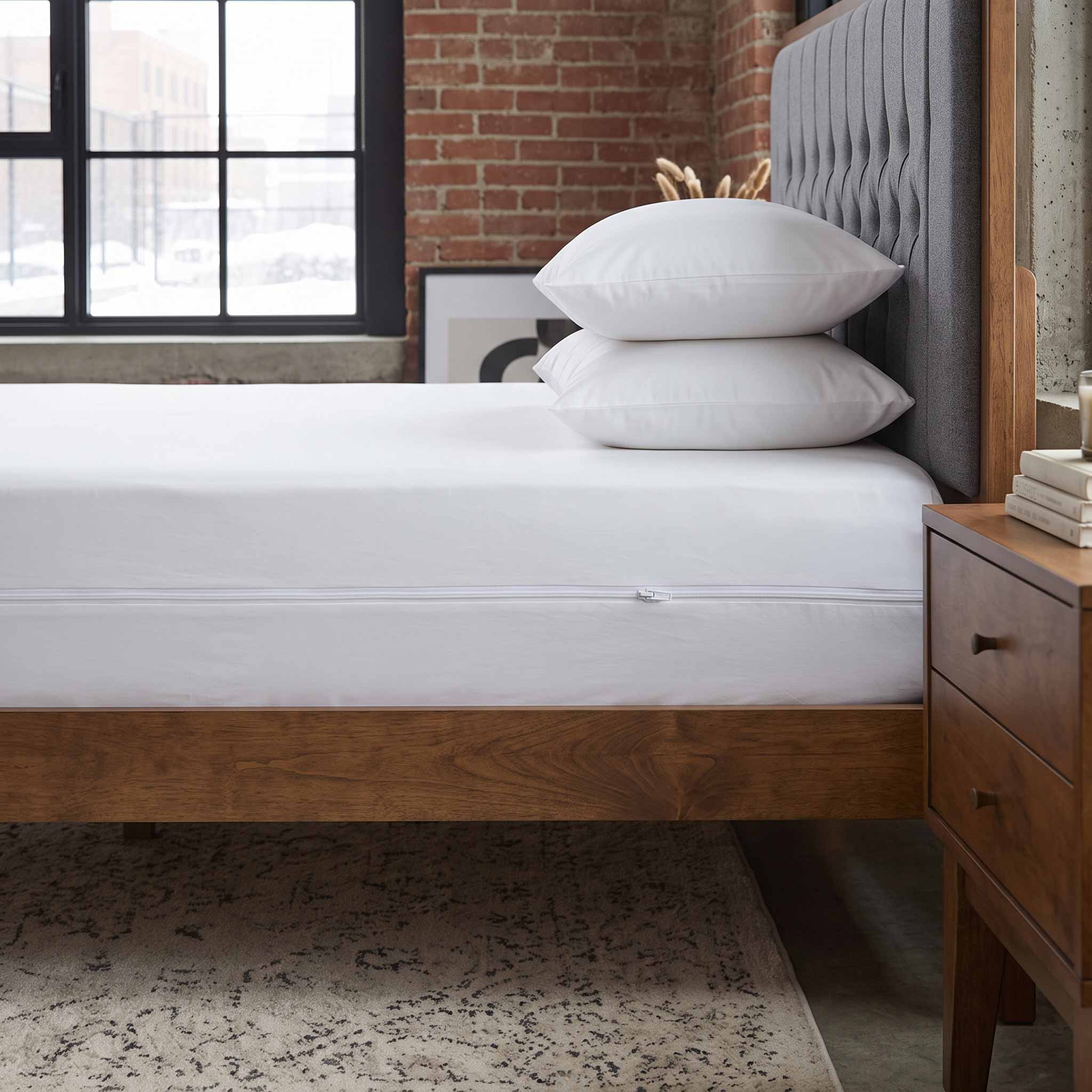 A white mattress fitted sheet and two stacked white pillows on a wooden bed frame with a gray tufted headboard, shown in a styled bedroom setting with a brick wall, window, and nightstand.