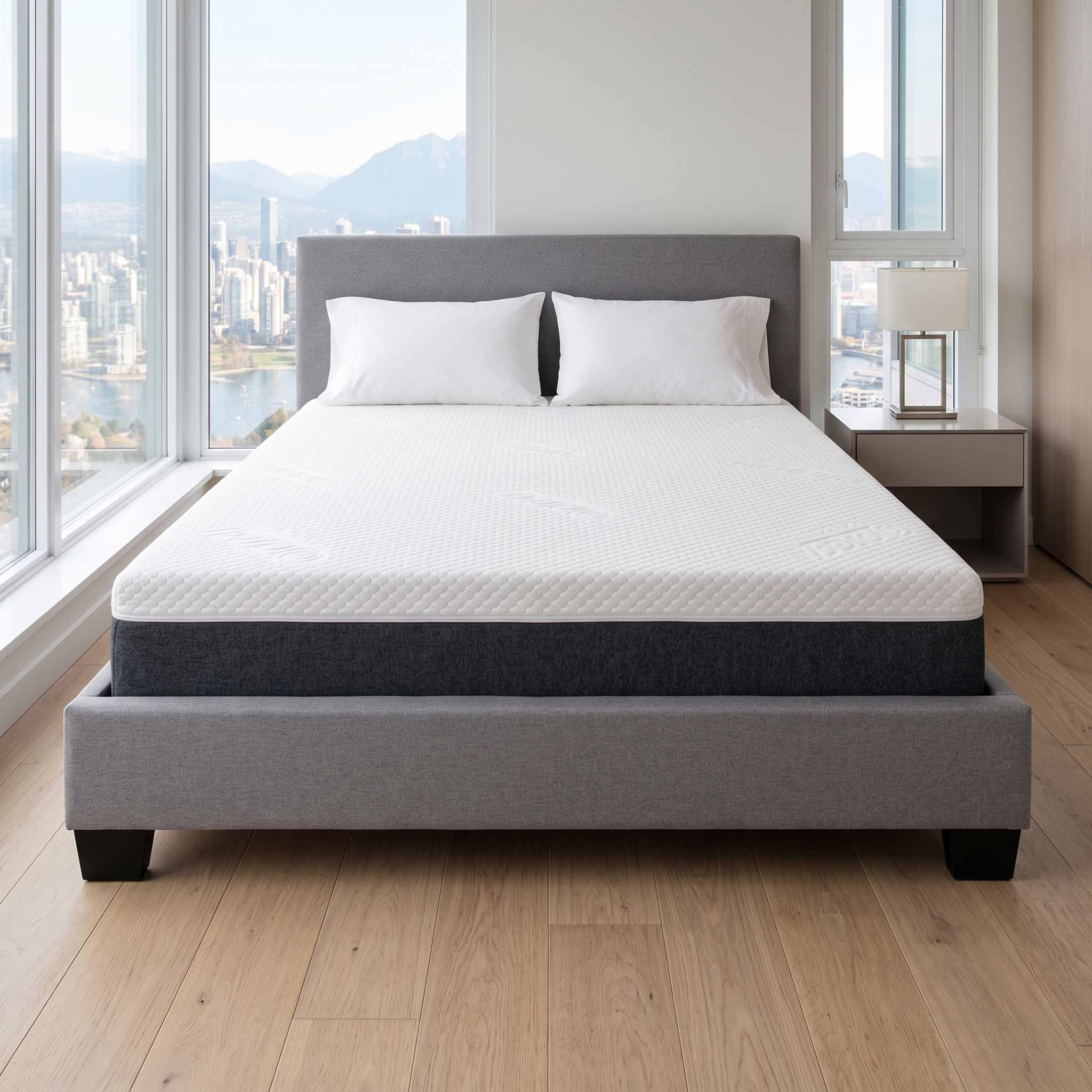A modern gray upholstered bed frame with a white quilted mattress and two white pillows, positioned in a bright bedroom with large windows overlooking a city skyline and mountains.