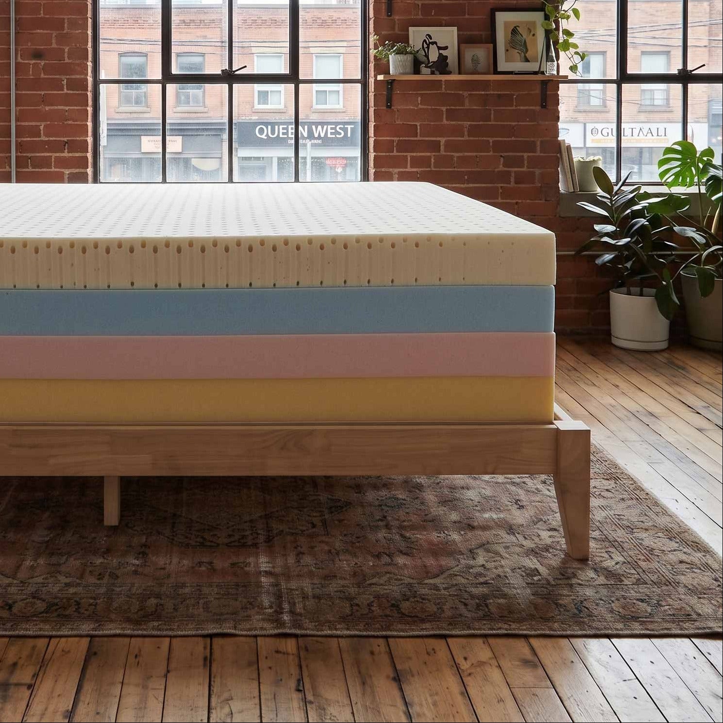 A high-profile mattress on a light wood bed frame, positioned in a loft-style bedroom with exposed brick walls, large industrial windows, and potted plants.