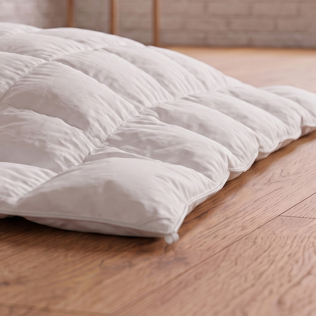 A white quilted down-style pillow or mattress topper resting on a light brown wooden floor in front of a softly blurred brick wall.