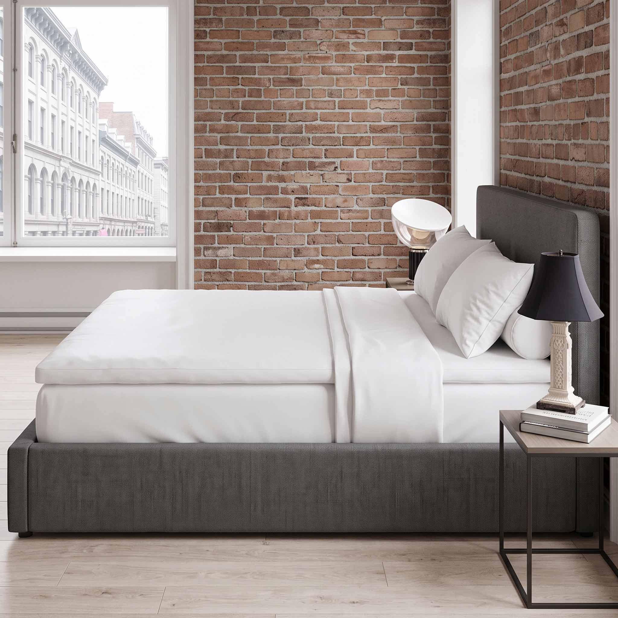A modern gray upholstered bed frame with white bedding and pillows, styled in a bedroom with a brick wall, window view, and a small black nightstand.