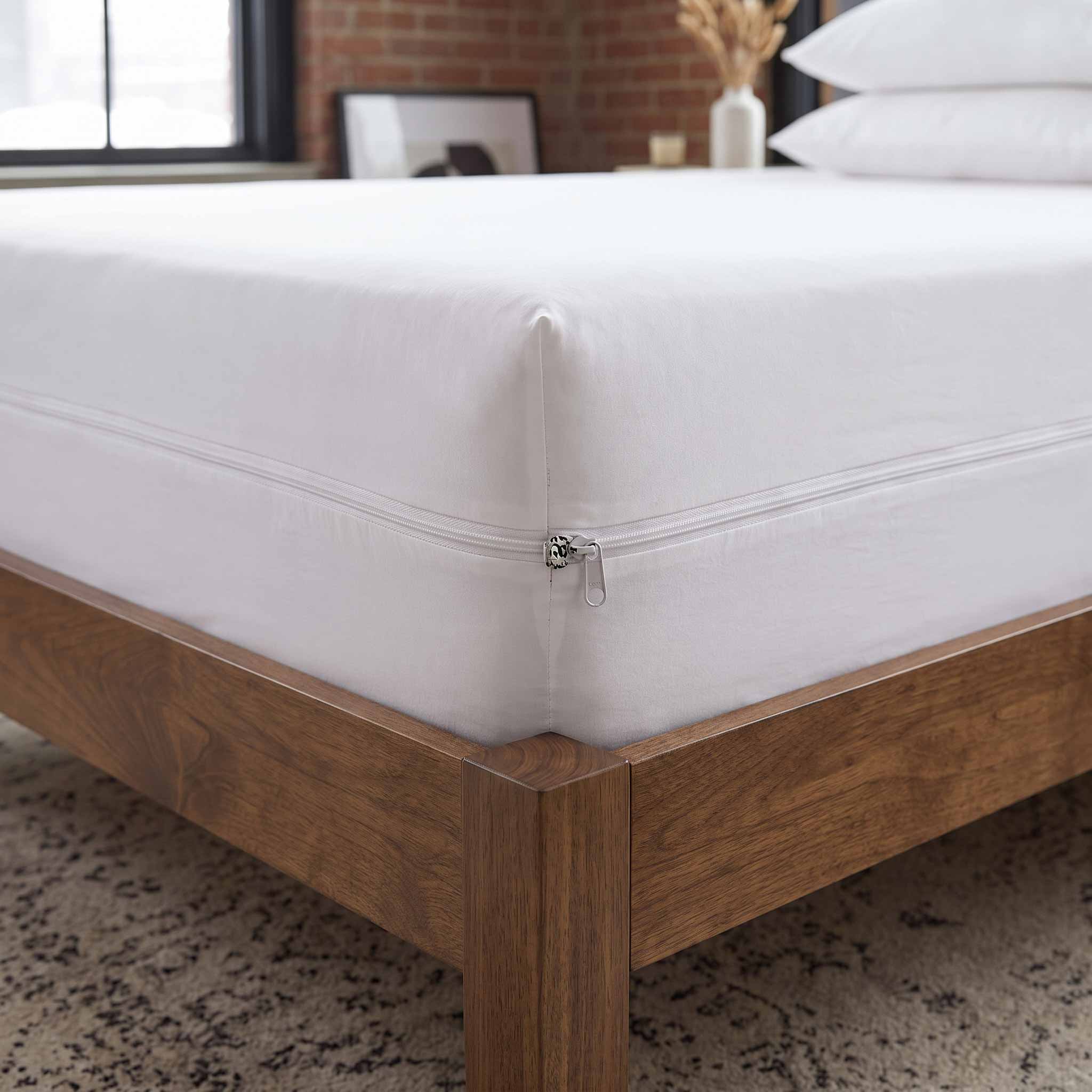 A close-up of a white mattress protector or fitted sheet wrapped around the corner of a wooden bed frame, with a zipper closure visible. The setting is a modern bedroom with a brick wall, window, and neutral rug.