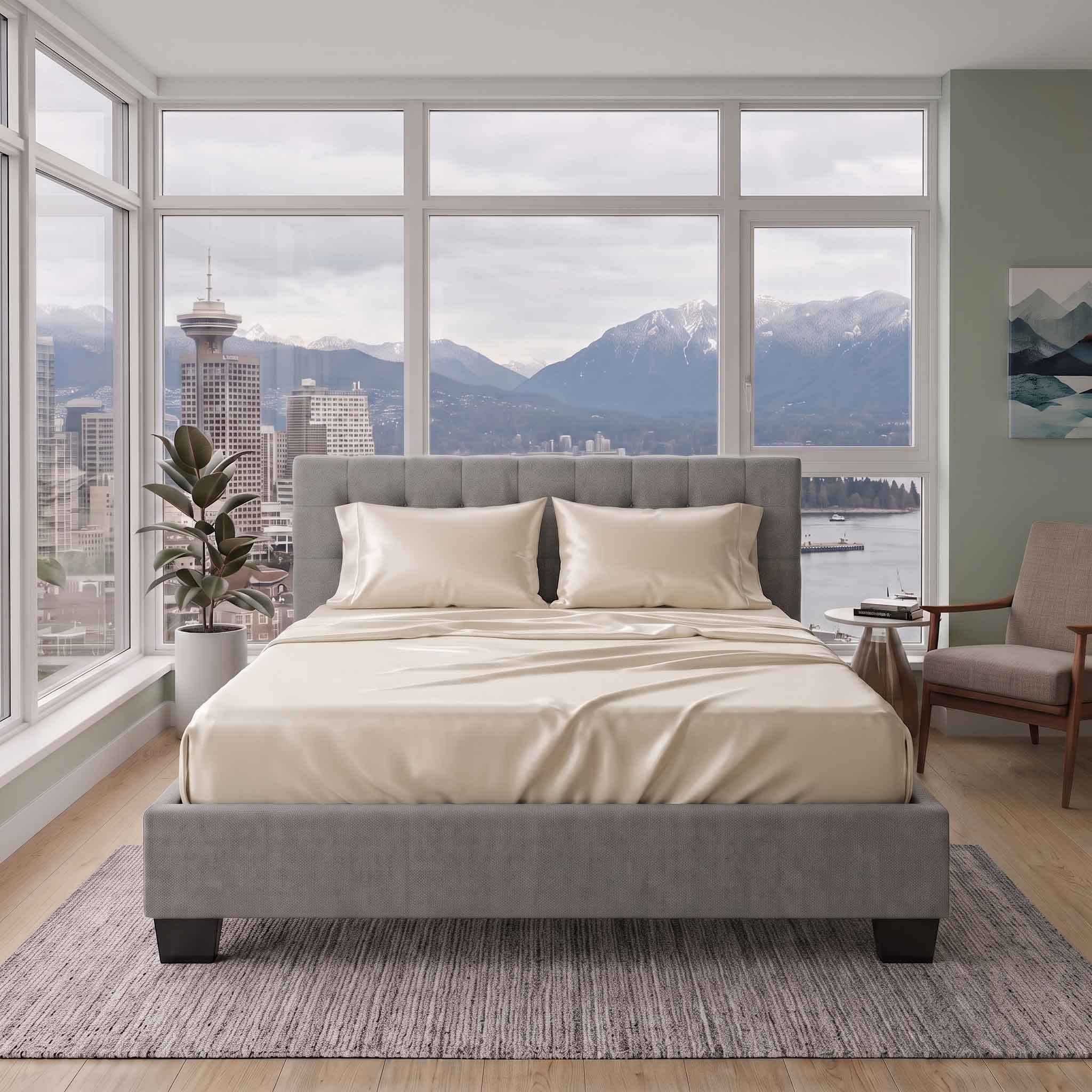 A gray upholstered bed with cream bedding is centered in a modern bedroom with large windows overlooking a city skyline and mountains. A matching gray upholstered chair sits to the right, and the room is styled with a rug, side table, and plant.