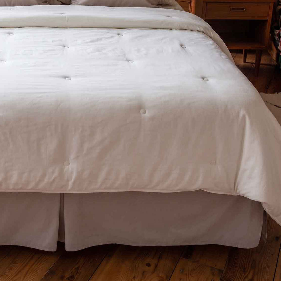 A neatly made bed in a bright bedroom, dressed with an off-white tufted comforter and matching pillows, set against a wooden headboard with large windows showing a scenic view of a lake and mountains.