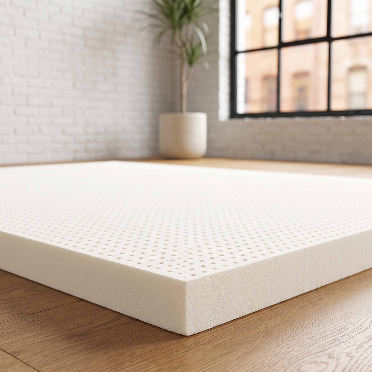 A thick, off-white latex mattress topper with a perforated surface pattern, shown resting on a light wood floor in a bright, modern room with a white brick wall and a window.