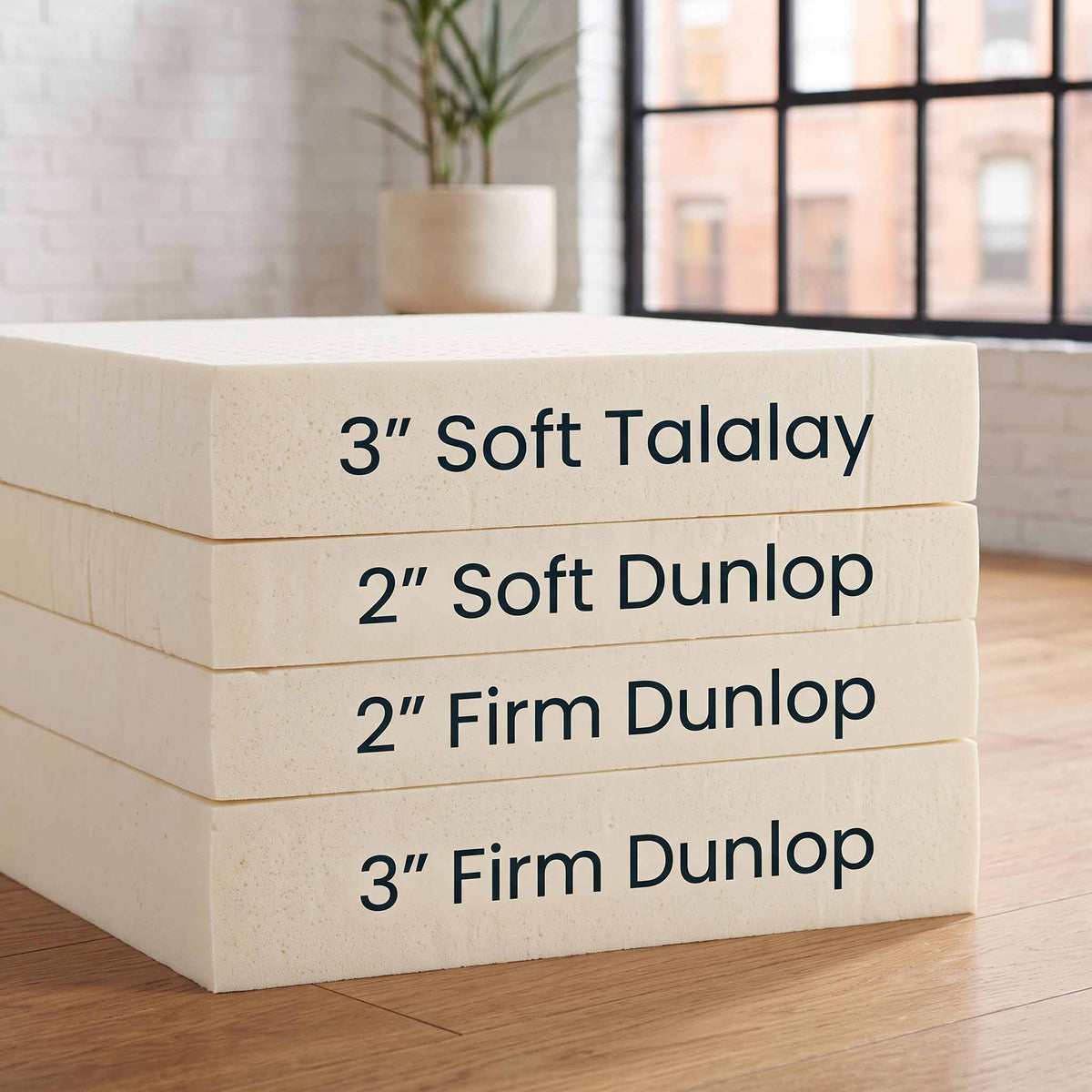 Four stacked foam mattress toppers on a wooden floor in front of a softly blurred window and brick wall. The top topper reads '3" Soft Talalay' and the lower two read '2" Soft Dunlop' and '2" Firm Dunlop'; the bottom topper reads '3" Firm Dunlop'.